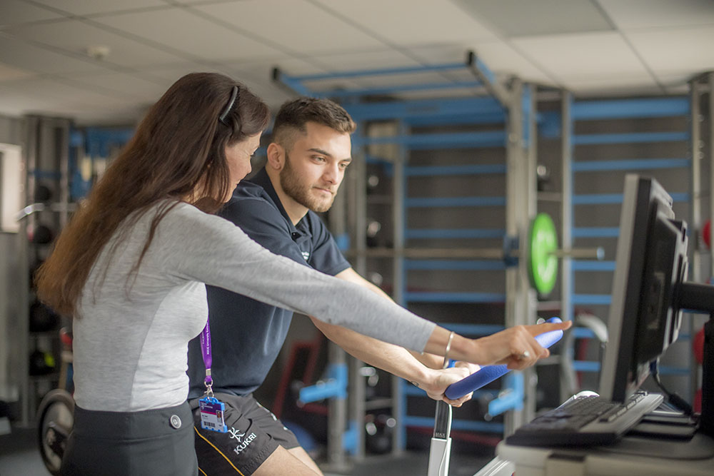A man rides an exercise bike whilst talking to a coach.