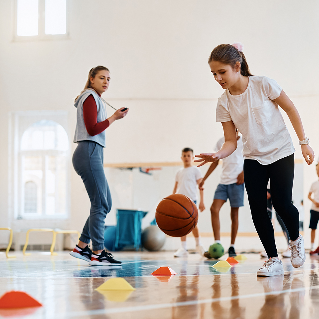 children leading a basketball during physical activity class at school gym. Her coach and friends are int he background.