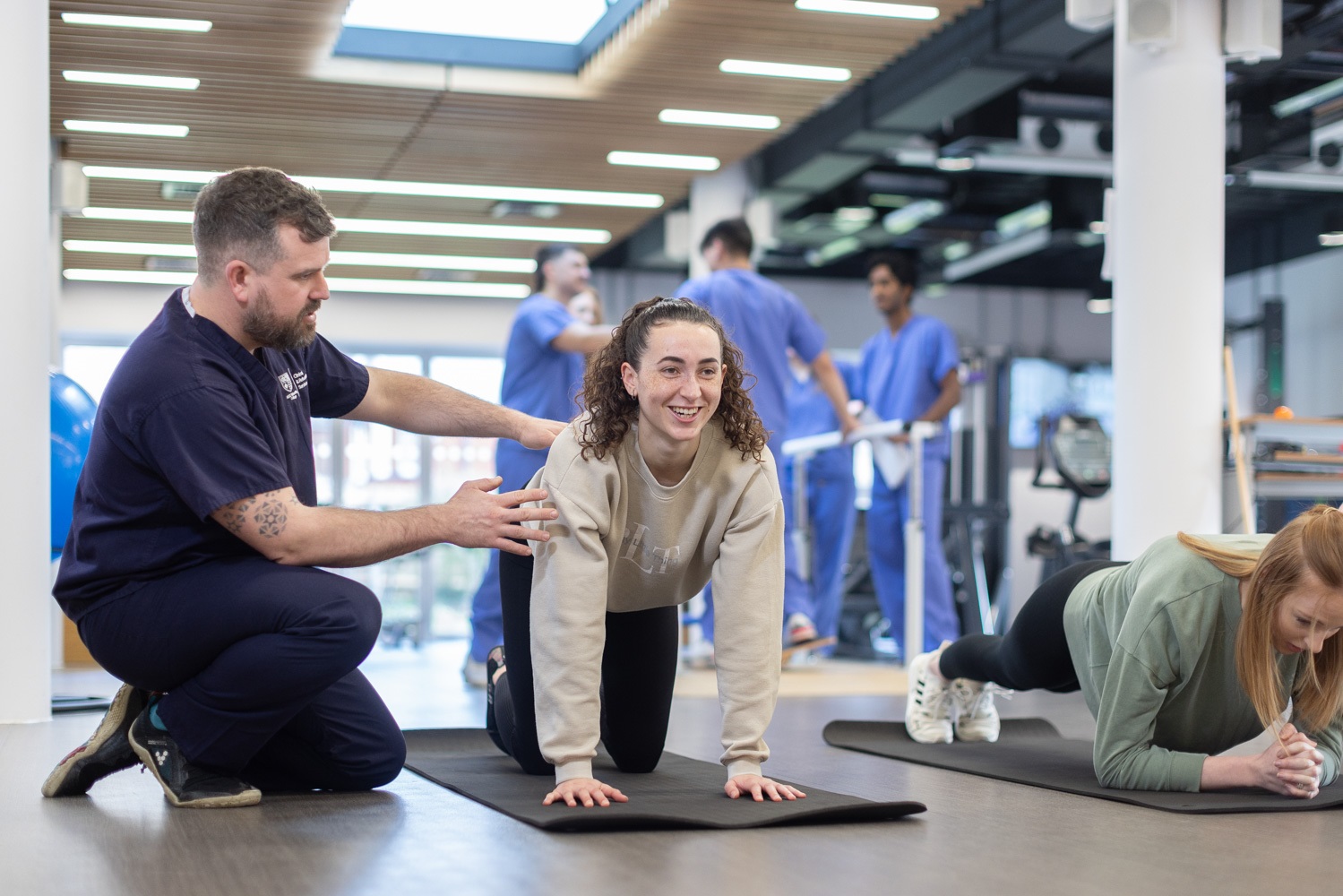 A woman does an excercise on a mat, whilst being coached,